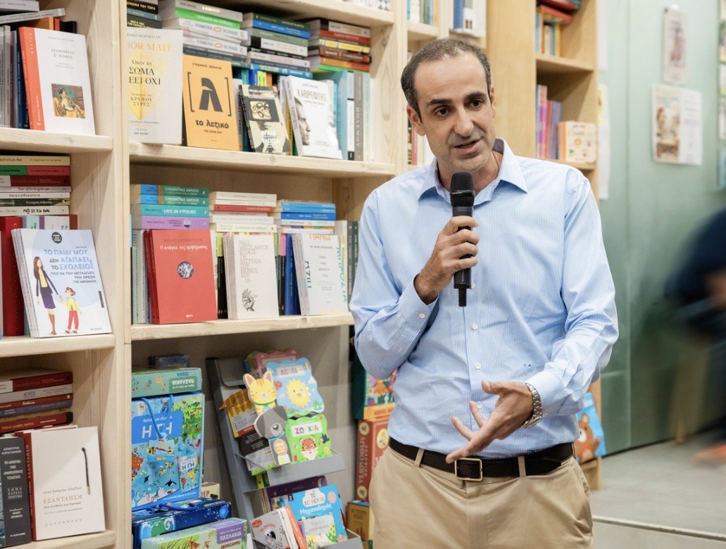 Ο Γρηγόρης Δημητριάδης μιλά με μικρόφωνο μπροστά σε βιβλιοθήκη